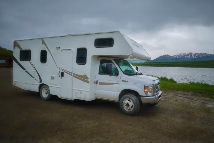 White RV rental parked on a dirt path beside a scenic lake with green trees and snow-capped mountains under a cloudy sky.. Thor Majestic 2018