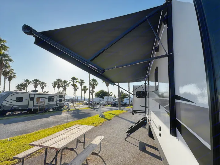 Enjoy your RV rental experience! This RV with its extended awning and picnic table is set up in a sunny park with palm trees.. Forest River Rockwood Ultra Lite 2024