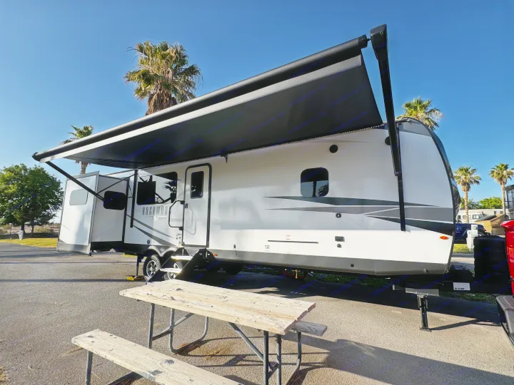 Sunny day with a white and grey RV rental set up at a campsite, featuring an extended awning and a picnic table.. Forest River Rockwood Ultra Lite 2024