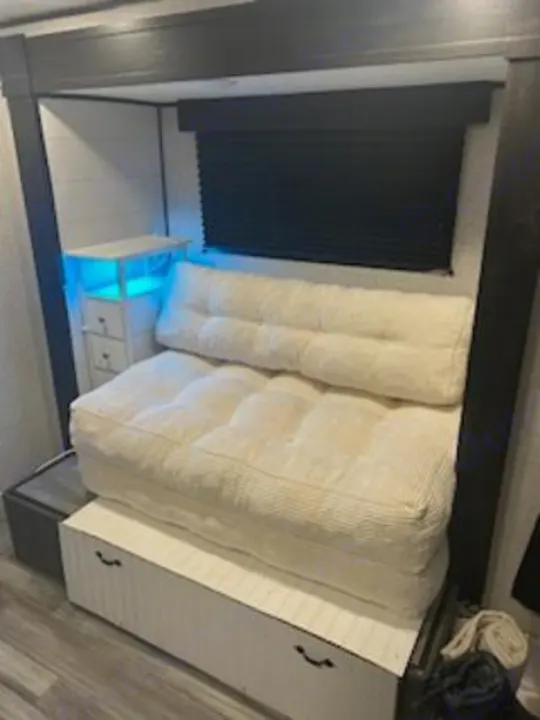 Compact RV rental space with a futon sofa over storage, illuminated side table, and a black window blind.. Jayco Jay Flight SLX 2024