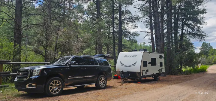 Black GMC Yukon SUV and Bullet travel trailer at a shaded forest campsite. Great RV rental for wilderness trips.. Keystone RV Bullet Crossfire 2021