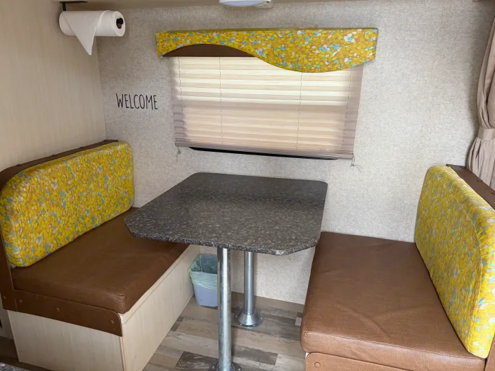 Cozy RV rental dinette with brown benches, colorful cushions, a dark table, window blinds, and a "WELCOME" sign on the wall.. Minnie Micro Minnie 2017