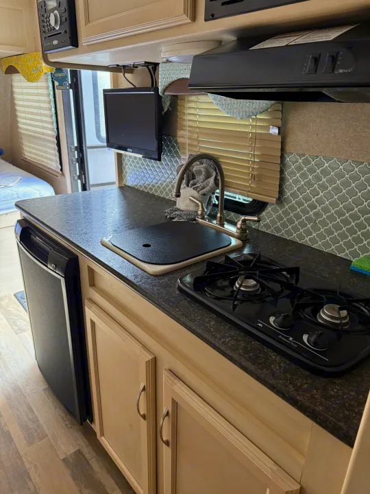 An RV rental kitchen with a dark countertop, sink, two-burner stove, small fridge, light cabinets, and green tiled backsplash.. Minnie Micro Minnie 2017