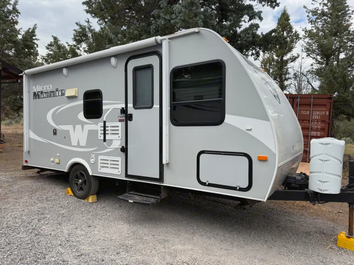 A grey Winnebago Micro Minnie travel trailer (rv rental) is parked on gravel, with trees and a shipping container behind it.. Minnie Micro Minnie 2017