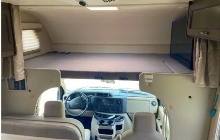 Spacious RV rental interior featuring a large cab-over bunk, front dashboard, and steering wheel. A TV is mounted right.. Coachmen Freedom Elite 2019