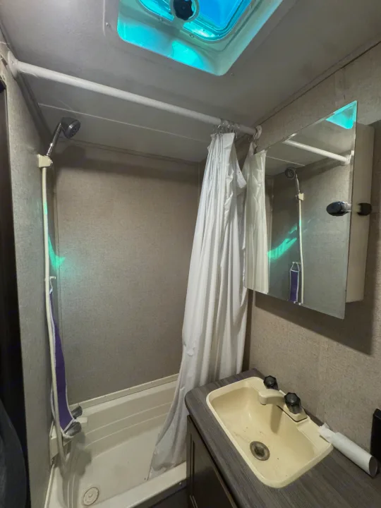 Compact RV rental bathroom featuring a white shower curtain, shower head, sink, and mirrored cabinet. A ceiling skylight adds light.. Dutchmen Coleman Lantern 2018