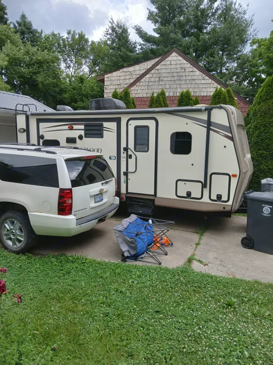 A large tan and black travel trailer, potentially an rv rental, parked on a concrete driveway with a white SUV. Lush trees and a house in the background.. Forest River Rockwood Ultra Lite 2016