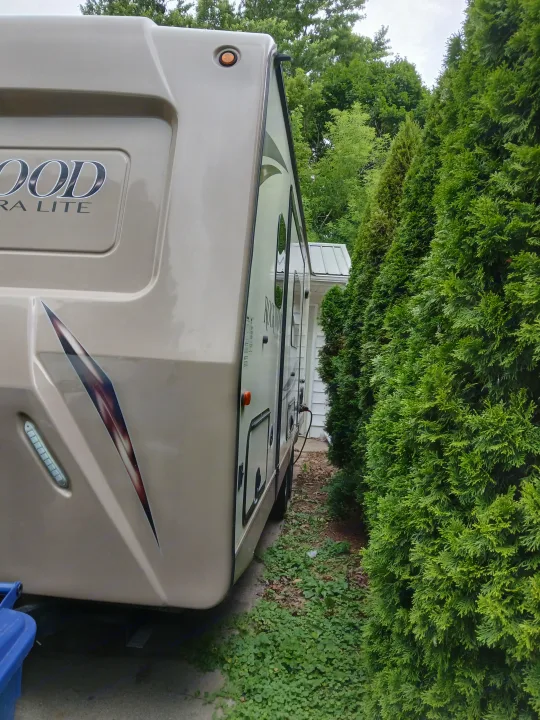 A beige Rockwood travel trailer RV rental, parked closely by tall green hedges & plugged into a house. Ready for use.. Forest River Rockwood Ultra Lite 2016