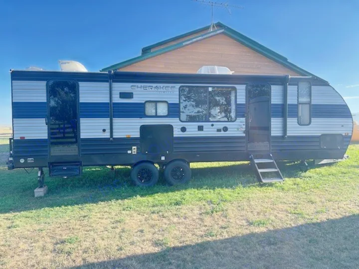 Side view of a blue & silver striped travel trailer, an rv rental, parked on green grass with a house and clear blue sky.. Forest River Cherokee 2022