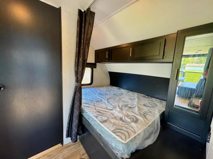 Compact RV rental bedroom featuring a new bed, dark overhead storage, a window, and a mirrored cabinet.. Forest River Grey Wolf 2021