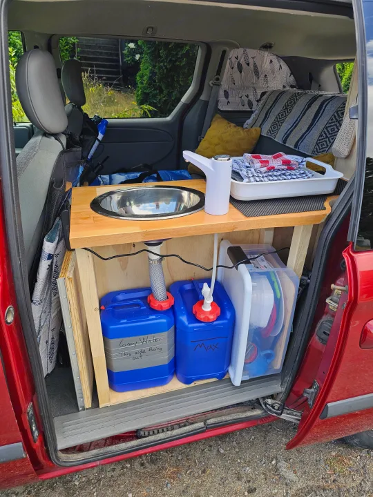Custom RV rental setup: a compact mobile kitchen in a van, featuring a wooden sink counter, electric faucet, and water storage tanks.. Dodge Caravan 2006