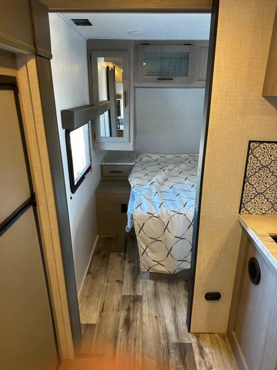 View into a modern RV rental bedroom, featuring a patterned bedspread, nightstand, overhead cabinet, and wood-look floor.. Coleman Dutchman Light 2023