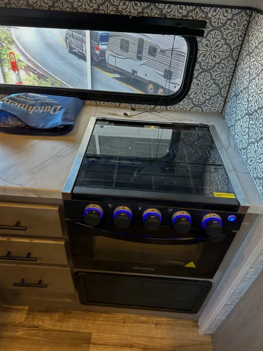 Inside a bright RV rental kitchen, featuring a GREYSTONE stove with blue-lit knobs, marble-patterned counters, and sleek walls.. Coleman Dutchman Light 2023