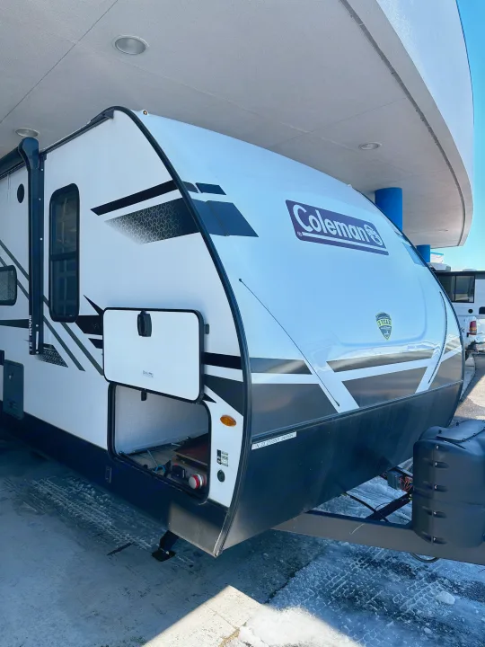 Front and side view of a white Coleman travel trailer, a great RV rental option, parked with an open storage hatch.. Coleman Dutchman Light 2023