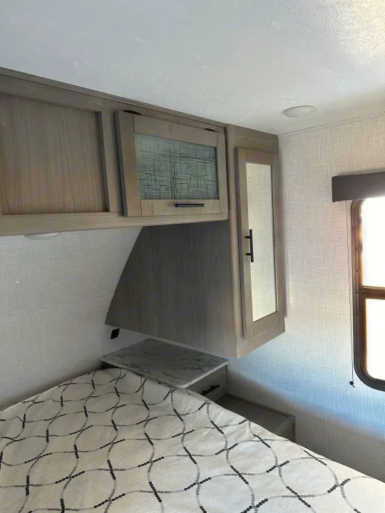 RV rental interior: view of a bedroom with overhead cabinets, patterned bed, and window.. Coleman Dutchman Light 2023