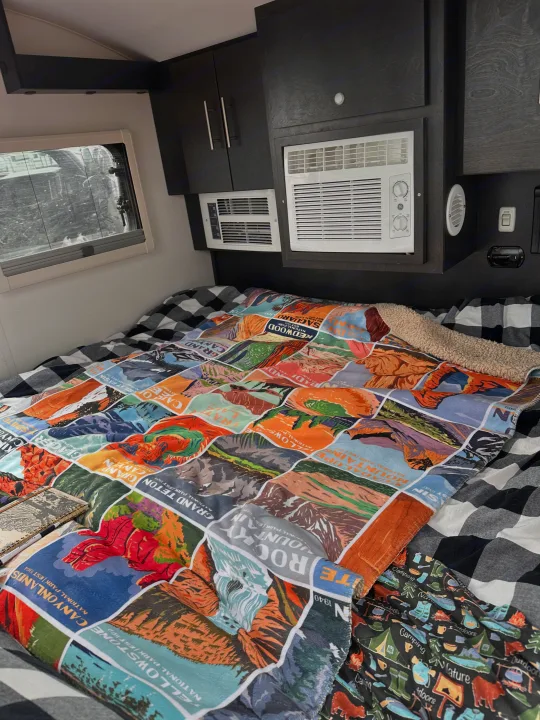 RV rental interior: cozy bed with buffalo check and National Park blanket, plus built-in AC units and dark cabinets.. nuCamp T@G 2023