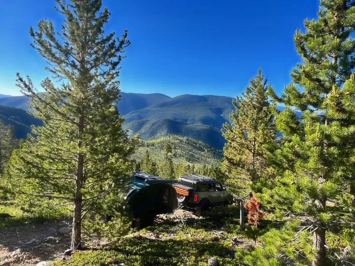 An off-road RV rental, a dark green Bronco towing a teardrop trailer, is parked among pine trees with sweeping mountain views.. Escapod Topo 2 2024
