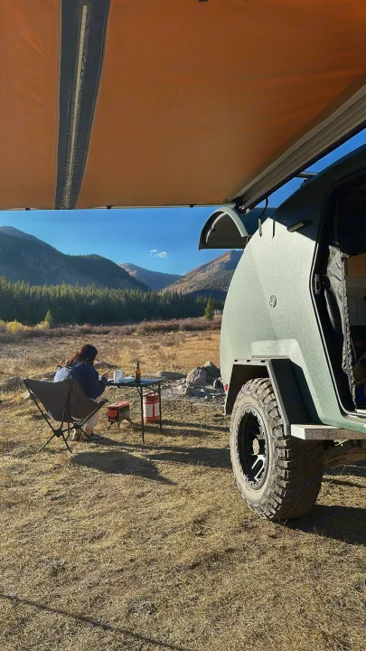 Camping with an RV rental: Person enjoys a meal beside a green teardrop trailer and awning in a mountain valley.. Escapod Topo 2 2024