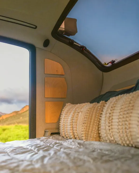 Inside a cozy RV rental: a comfortable bed, wooden shelves, and a large skylight showing blue sky, with a scenic outdoor view.. Escapod Topo 2 2024
