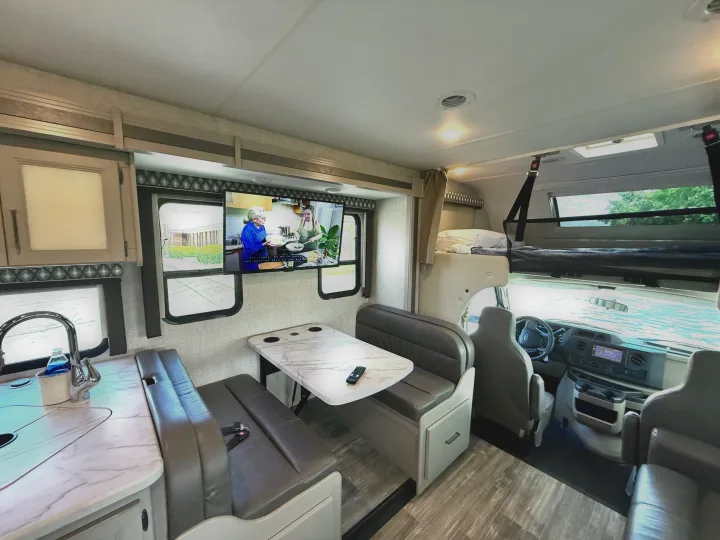 An RV rental's inviting interior, showing the dinette, galley kitchen, TV, and overhead bunk. Ready for your trip!. Thor Geneva 2022