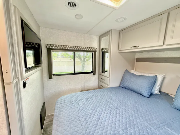 Bright RV rental bedroom featuring a bed with blue quilted bedding, window, TV, and built-in storage.. Thor Geneva 2022