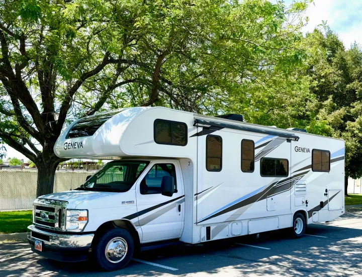 A white Geneva E450 Class C rv rental parked in a sunlit lot with green trees. Perfect for your next adventure!. Thor Geneva 2022