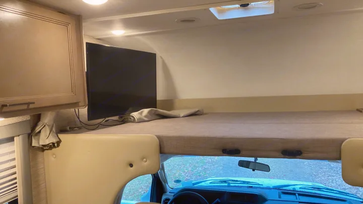 Inside an rv rental: a cozy cab-over bunk with a bed, TV, and overhead storage, above the driver's cabin.. Thor Four Winds 2022