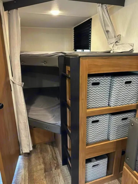 RV rental interior: cozy bunk beds with privacy curtains, a ladder, storage bins, and wood-look flooring.. Forest River Campsite Ultra 26CJL 2025