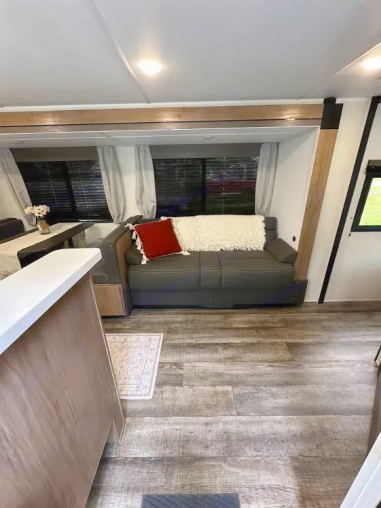 Cozy RV rental interior with a dark sofa, red and white pillows, and wood-look floor. Dinette & kitchen island visible.. Forest River Campsite Ultra 26CJL 2025