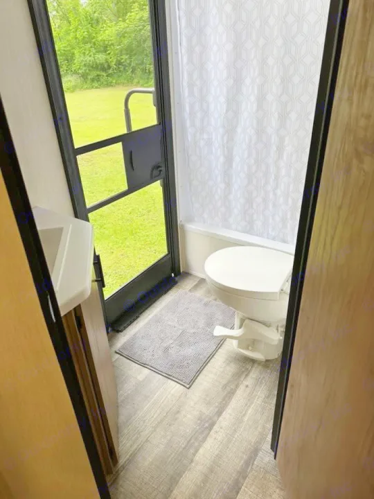 Inside an RV rental: a compact bathroom featuring a toilet, shower curtain, sink, and outdoor view through the screen door.. Forest River Campsite Ultra 26CJL 2025