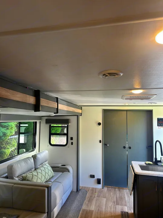 Clean RV rental interior featuring a light grey sofa, a small kitchen area with a sink, and dual doors, offering a homey feel.. Keystone RV Bullet Crossfire 2024