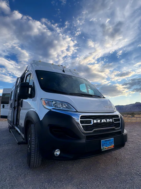 A white Ram RV rental van, parked on a gravel lot under a dramatic cloudy sky, with mountains in the background.. Winnebago Winnebago Solis 59PX Dodge Ram 2023
