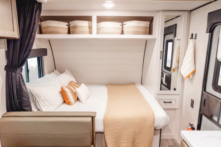 Cozy RV rental bedroom featuring a neatly made bed, decorative pillows, overhead storage with baskets, and a window with dark curtains.. Wayfinder RV Go Play 19BH 2025
