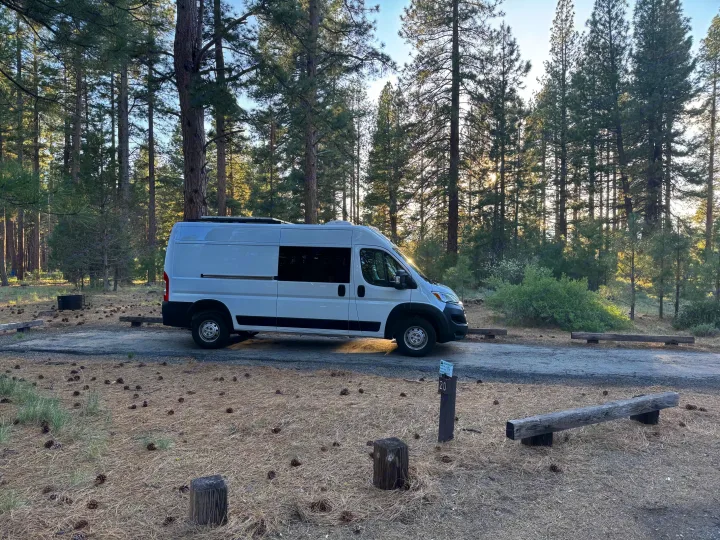 White camper van at campsite 20 amidst pine trees. Perfect RV rental for nature getaways.. Dodge B Van 2023
