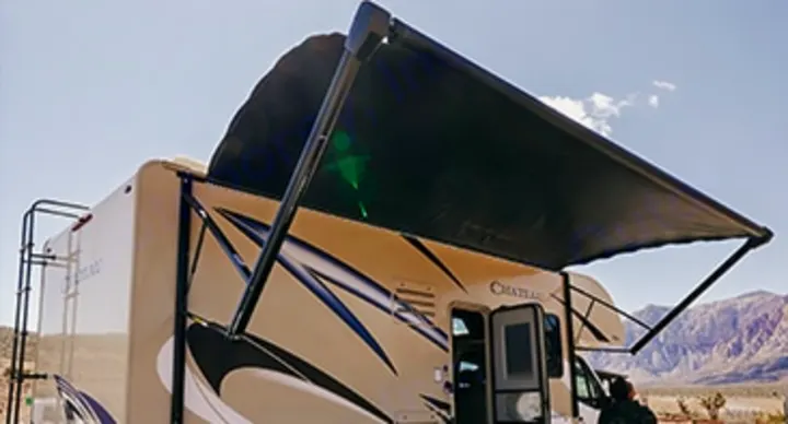 A light-colored RV rental has its dark awning extended, casting shade over a desert landscape with mountains. Ready for outdoor adventure.. Thor Chateau Sprinter 2020