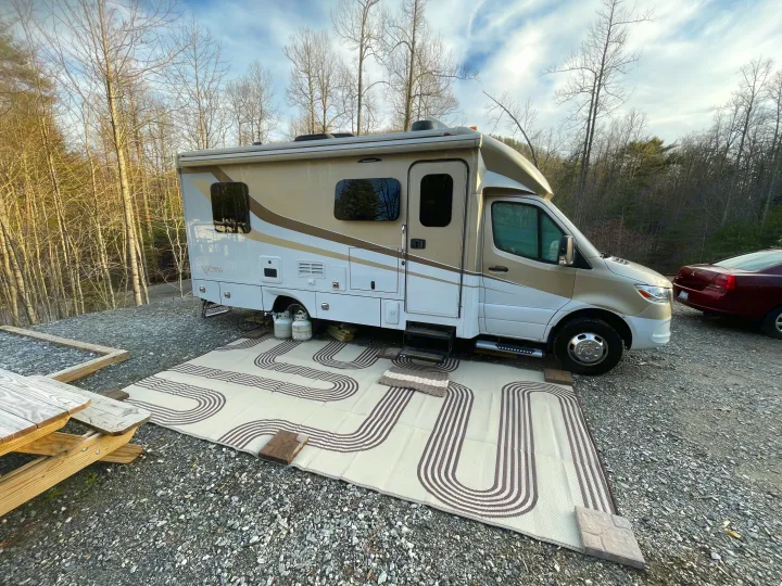 A beige & white RV rental parked on a gravel campsite. An outdoor mat, picnic table, & bare trees complete the peaceful scene.. Renegade Custom Coaches Vienna 2021