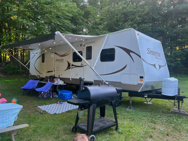 Cozy RV rental setup with an extended awning, string lights, and a BBQ grill in a grassy, wooded campsite.. Keystone RV Sprinter Select 2010