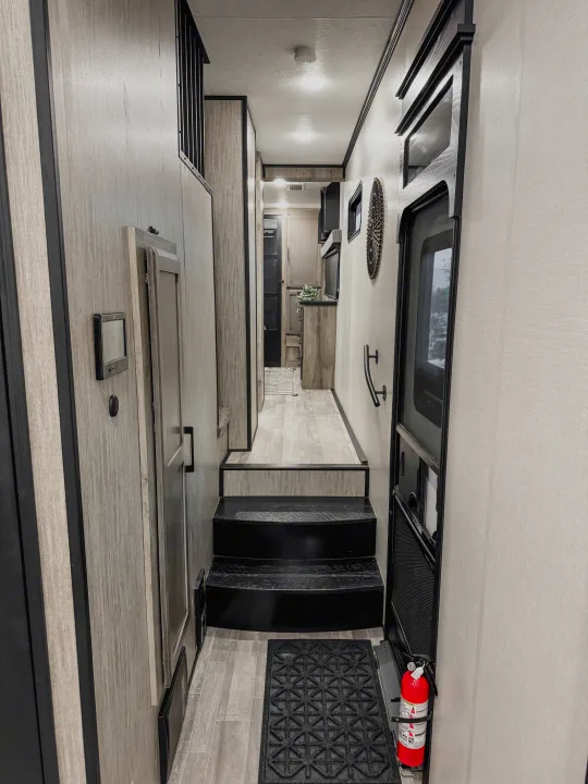 Explore this RV rental's modern interior: dark stairs lead to a light wood-grain hallway. Features include a fire extinguisher and kitchen view.. Durango Gold 2023