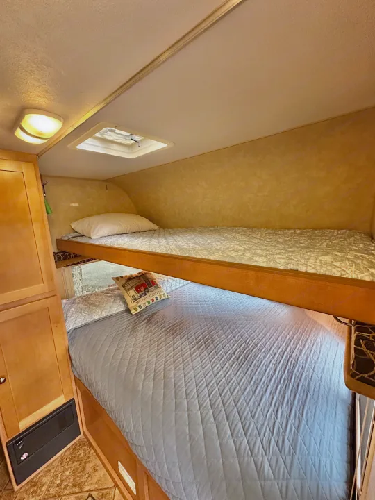 Cozy RV rental interior with two bunk beds. Features light blue and white quilted bedding, a window, and wood cabinetry.. Cruiser RV Fun Finder X 2011