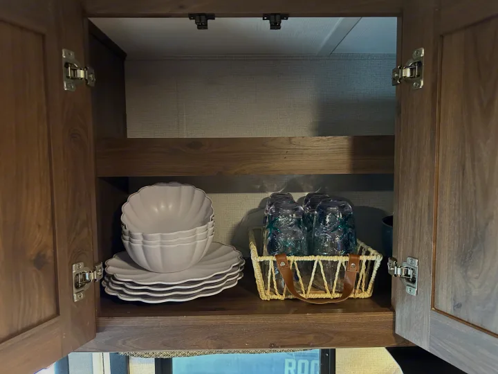 Organized RV rental cabinet with light grey scalloped dishes and blue textured glasses in a basket.. Forest River Sunseeker LE 2021