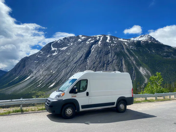 An RV rental (Ram Promaster) parked on a mountain road with towering, snow-dusted peaks and a blue sky.. Ram Promaster 2014