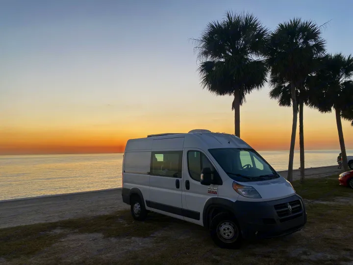 Enjoy van life! White rv rental parked by the ocean shore at sunset, with silhouetted palm trees.. Ram Promaster 2014