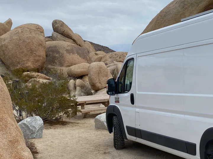 A white RV rental camper van parked amidst giant boulders and desert plants with a picnic table in a rugged landscape.. Ram Promaster 2014