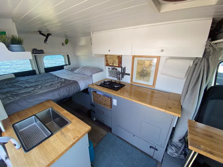 Interior of a well-equipped RV rental camper van featuring a bed by ocean-view windows & a functional kitchenette.. Ram Promaster 2014