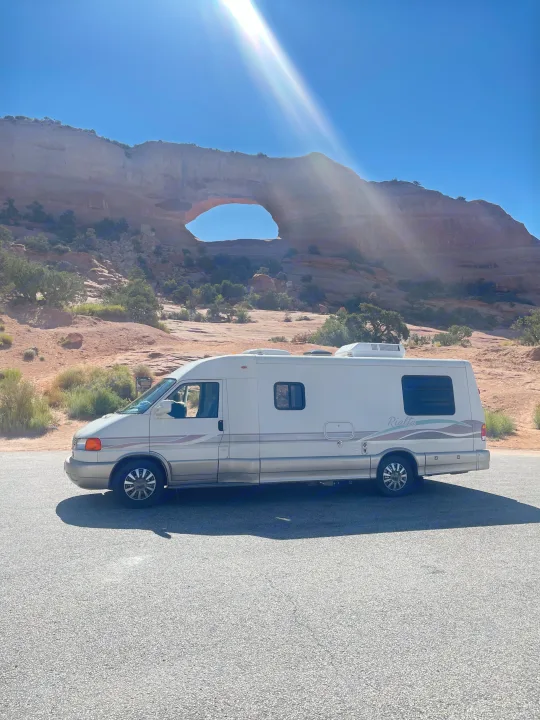 White RV rental parked at a majestic desert natural arch under a bright blue sky, perfect for a scenic road trip.. Eurovan Camper Rialta 2000