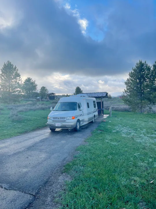 A white RV rental is parked on a gravel path at a cloudy campground, surrounded by green grass and pine trees.. Eurovan Camper Rialta 2000