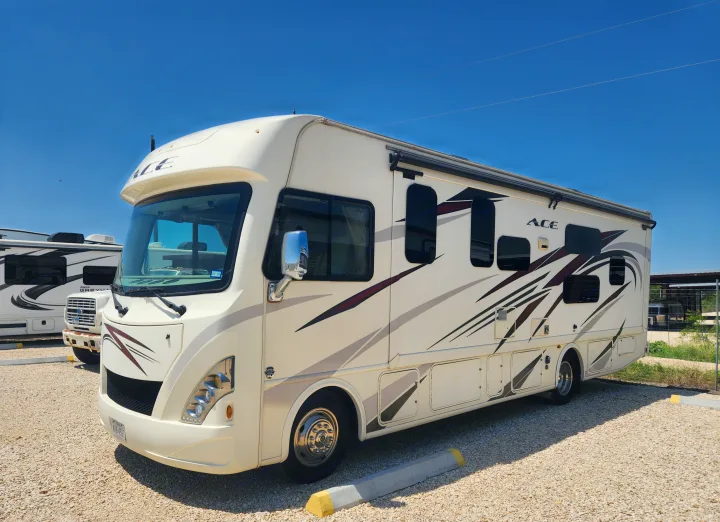 A cream-colored ACE rv rental, a Class A motorhome with dark decals, parked on gravel under a bright blue sky.. Thor Motor Coach ACE 2018