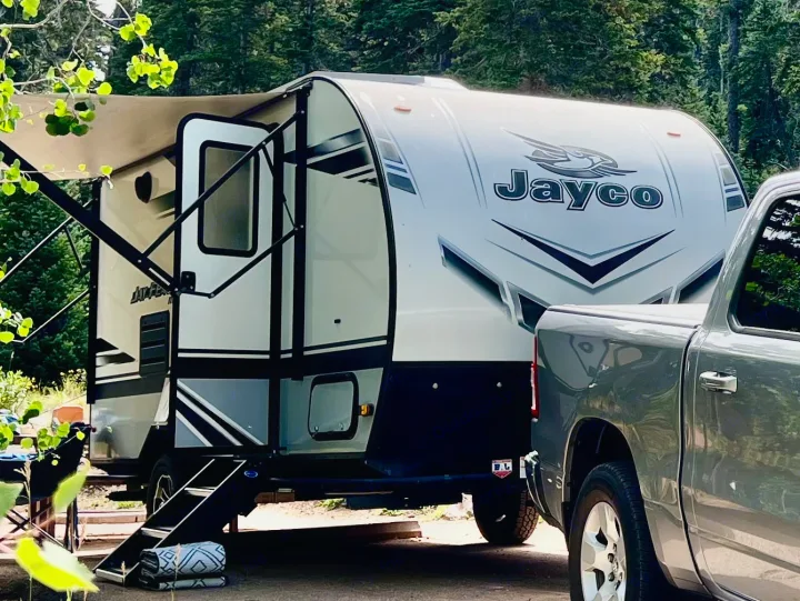 This Jayco RV rental, a travel trailer with an extended awning and steps, is parked with a gray pickup truck at a wooded campsite.. Jayco Jay Feather Micro 2021