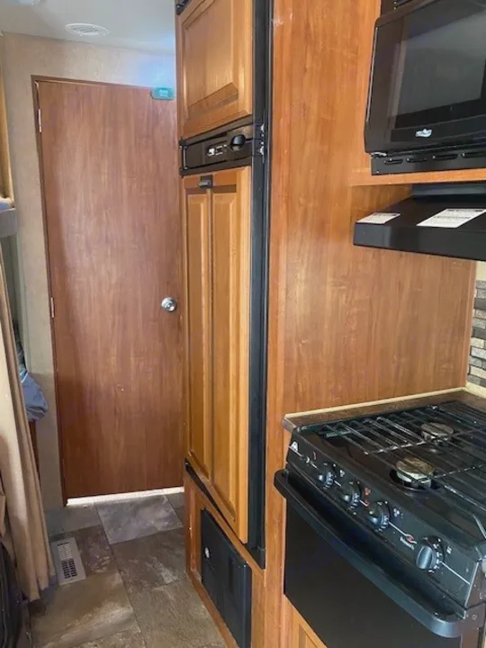 This RV rental interior shows a compact kitchen with a stove, microwave, and refrigerator, plus a wooden door.. Jayco Jay Flight 2013