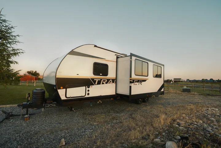 Modern white and black RV rental parked on a gravel lot with a slide-out extended, surrounded by fields and a fence.. Prime Time Tracer 270BHSLE 2024